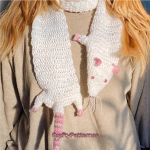 May include: A white crochet scarf with a rat design. The scarf features a textured stitch, pink accents on the paws, ears, and tail, and small red bead eyes. The tail has pink and white stripes. The scarf is handmade.