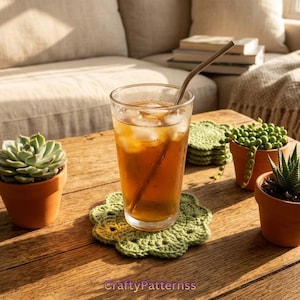 May include: A clear glass filled with amber liquid, ice cubes, and a metal straw, resting on a light green crocheted flower-shaped coaster. Two terracotta pots containing a succulent and a string of pearls plant sit nearby on a wooden surface.