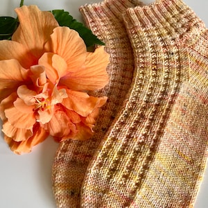 Könnte beinhalten: Ein Paar handgestrickte Socken in Gelb-, Rosa- und Brauntönen mit Rippenmuster am Bein. Eine große, pfirsichfarbene Hibiskusblüte mit grünen Blättern liegt neben den Socken. Die Socken sind aus weichem Garn gefertigt.