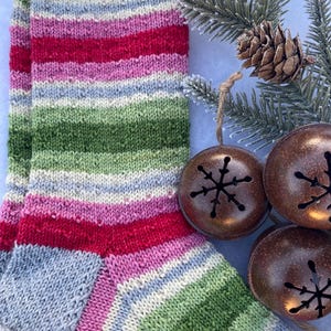 Könnte beinhalten: Gestreifte Stricksocken in den Farben Grün, Rosa, Rot und Blau. Die Socken liegen auf einer weißen Oberfläche mit einem Tannenzweig und dekorativen Glocken.