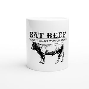 Op de afbeelding: Een witte keramische mok met een zwart-witte illustratie van een koe en de tekst "Eat Beef The West Wasn't Won On Salads".