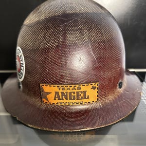 May include: A brown hard hat with a textured surface. The hat features a patch that reads "TEXAS ANGEL" in gold lettering, with a star and Texas outline. A circular sticker is also visible.