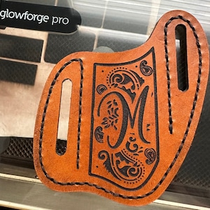 May include: A brown leather knife sheath with a decorative black engraved design featuring the letter "M" and floral and paisley patterns. The sheath has two slots and is stitched with black thread. The text "glowforge pro" is in the upper left corner.