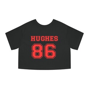 Op de afbeelding: Zwart gecropped T-shirt met rode tekst "HUGHES 86" in een varsity-letter-stijl.