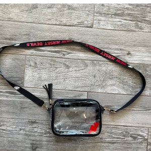 May include: Clear plastic crossbody bag with a black strap and a black tassel. The strap has the words "NEW JERSEY DEVILS" printed in red.