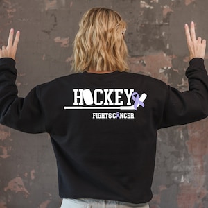 Puede incluir: Sudadera negra con texto blanco que dice "Hockey Fights Cancer" y una cinta morada.