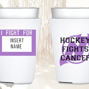 Puede incluir: Dos portavasos blancos aislados. Uno tiene el texto "I Fight For" con un espacio en blanco para un nombre. El otro tiene el texto "Hockey Fights Cancer" con un logotipo de palo de hockey morado.