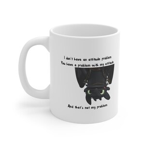 May include: White ceramic coffee mug featuring a black and white illustration of a dragon hanging upside down. The text on the mug reads: "I don't have an attitude problem. You have a problem with my attitude. And that's not my problem."