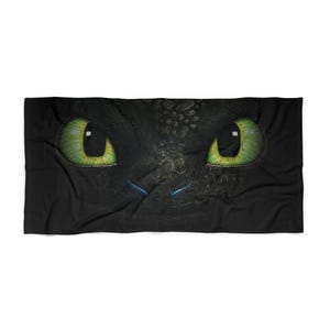 May include: Black rectangular blanket featuring a close-up of a dragon's face. The dragon has large, bright green eyes with yellow highlights. The skin is black with a textured pattern, and there are blue accents near the nose.