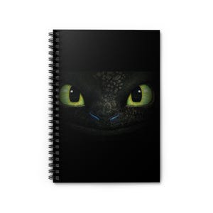 Può includere: Un quaderno nero con rilegatura a spirale. La copertina presenta un primo piano del viso di un drago con occhi verdi.