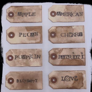 May include: A collection of ten vintage-style paper tags with brown stains and a metal eyelet. Each tag features a word in black ink: APPLE, AMERICAN, PECAN, CHERRY, PUMPKIN, PATRIOTIC, BLUEberry, LOVE, PEACH, and LEMON.