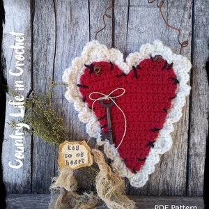 May include: A crocheted red heart with a white crocheted border, a key, and a tag that says "key to my heart". The heart is hanging on a rustic wooden background.
