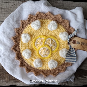 May include: Crochet pattern for a lemon meringue pie, featuring a yellow and white crocheted pie with a brown crust. The pie is on a white cloth with a tag that says "Sunshine".