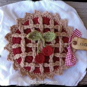 Puede incluir: Tarta de cerezas de crochet con una corteza roja y marrón, hojas verdes y cerezas rojas. La tarta está sobre un paño blanco con una cinta a cuadros rojos y blancos y una etiqueta marrón que dice "CHERRY".