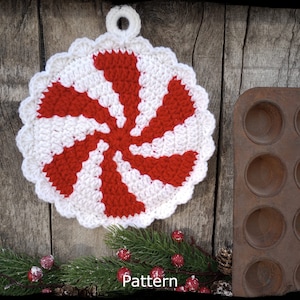 May include: A red and white crocheted peppermint candy pot holder with a loop for hanging. The pot holder is on a wooden background with a metal muffin tin and a sprig of evergreen.