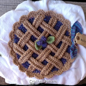 Puede incluir: Patrón de ganchillo para una fuente de pastel de arándanos. La fuente está hecha con hilo marrón y morado y tiene un borde festoneado. La fuente está decorada con un arándano y hojas de ganchillo.