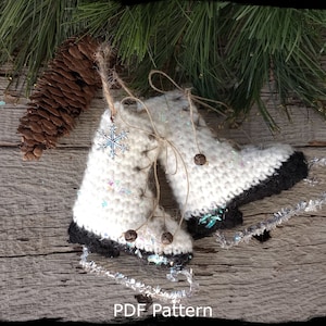 May include: Crochet ice skate ornaments with white yarn, black trim, and silver bells. The ornaments are on a rustic wooden surface with a pine branch and pine cone in the background. The text "Country Life in Crochet" and "PDF Pattern" are visible in the image.