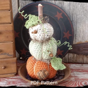 Puede incluir: Una decoración de calabaza de ganchillo apilada en tonos naranja, verde y crema, con un tallo de ramita y acentos de vid verde. La calabaza se exhibe en un cuenco de madera. El texto "Country Life in Crochet" y "PDF Pattern" son visibles.