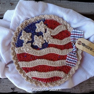 May include: Crocheted American flag design with red, white, and blue yarn. The flag is round and has a crocheted border. The flag is on a white cloth with a red, white, and blue checkered ribbon and a brown tag that says "AMERICAN".