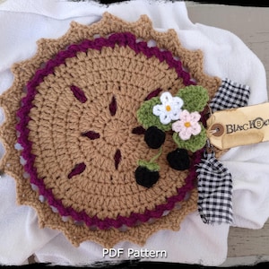 Peut inclure: Plat à tarte crocheté avec une bordure violette et un centre brun. Le plat à tarte est décoré de fleurs et de baies crochetées. Le plat est sur un fond blanc avec une étiquette qui dit "BlackBerry".