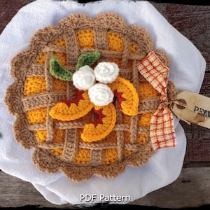 Peut inclure: Plat à tarte en crochet en forme de pêche avec une croûte brune, une garniture de pêche jaune et des fleurs blanches en crochet. Le plat est sur un fond blanc avec un ruban à carreaux rouge et blanc et une étiquette brune qui dit "PEACH".