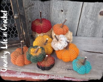Puede incluir: Una colección de calabazas de ganchillo en varios colores, incluyendo naranja, rojo, verde y azul. Las calabazas están dispuestas sobre una superficie de madera y tienen pequeñas ramitas unidas a las tapas. El texto "Country Life in Crochet" es visible en el lado izquierdo de la imagen. El texto "PDF Pattern" es visible en la parte inferior de la imagen.