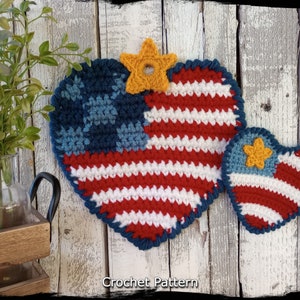 May include: Two crocheted American flag hearts, one large and one small, with a yellow star on each. The hearts are hanging on a white wooden background. "Country Life in Crochet" and "Crochet Pattern" are written below the hearts.