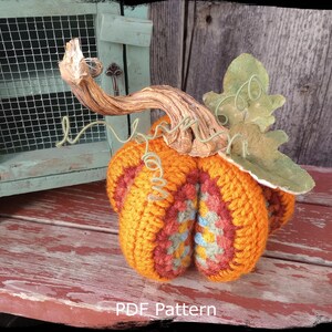 May include: Crocheted pumpkin with a brown twig stem and a green leaf. The pumpkin is orange, red, and yellow with a blue and green pattern. The pumpkin is sitting on a red wooden surface. PDF Pattern.