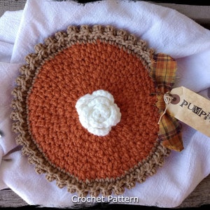 Peut inclure: Tarte à la citrouille en crochet avec une fleur blanche en crochet sur le dessus. La tarte est sur une serviette blanche avec un tissu à carreaux et une étiquette qui dit "PUMPKIN".