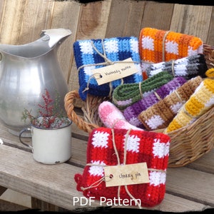 May include: A rustic scene featuring a woven basket filled with colorful, crocheted items. The items, including washcloths and scrubbies, are in various colors like blue, orange, and red, with white accents. Each item is tied with twine and labeled. A metal pitcher and mug are also present.