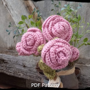 May include: A crocheted bouquet of pink roses in a rustic metal container with a burlap bow and a tag that says "Bloom". The roses are arranged in a vase with green leaves. The background is a weathered wooden surface.