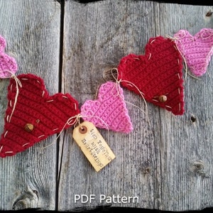 Puede incluir: Una guirnalda de corazones de crochet con corazones rojos y rosas alternados. La guirnalda está enhebrada con hilo y tiene pequeñas campanas unidas a cada corazón. Una etiqueta está unida a uno de los corazones con el texto "Tied Together with Heartstrings".
