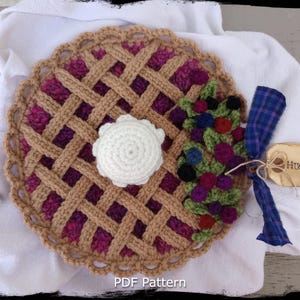 May include: Crocheted pie with a white crocheted dollop on top and a cluster of purple, green, and blue crocheted berries on the side. The pie is on a white fabric background with the text "Country Life in Crochet" and "PDF Pattern".