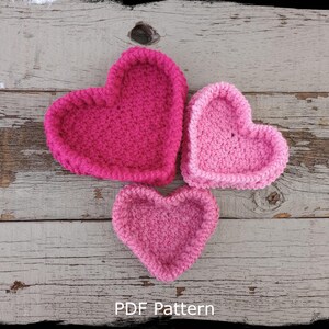Może przedstawiać: Trzy plecione koszyki w kształcie serca w odcieniach różowego i fuksji, ułożone na powierzchni z postarzanego drewna. Tekst "Country Life in Crochet" i "PDF Pattern" jest widoczny pod koszykami.