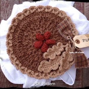 Peut inclure: Un motif de tarte aux noix de pécan crochetée, avec une croûte et une garniture marron, des accents décoratifs de fleurs et de feuilles. Une étiquette avec l'inscription "Pecan" est attachée. Les mots "Country Life in Crochet" et "PDF Pattern" sont également visibles.
