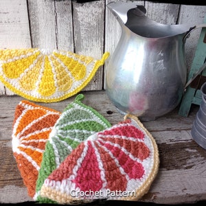May include: Four crocheted lemon slice hot pads in yellow, orange, green, and red. The hot pads are arranged on a wooden surface with a silver jug and metal containers in the background. The text "Country Life in Crochet" is written vertically on the left side of the image. The text "Crochet Pattern" is written horizontally at the bottom of the image.