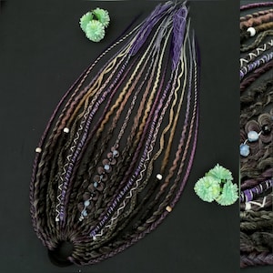 May include: A collection of synthetic dreadlocks in shades of purple, brown, and beige, with decorative beads and white stitching. The dreadlocks are arranged on a black surface, with small green decorative accents.
