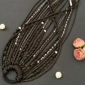 May include: Dark brown dreadlocks with white accents and small white beads. The dreadlocks are styled with a black elastic band. The image also includes small white and pink flowers, creating a contrast against the dark hair.