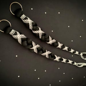 May include: A pair of black braided hair extensions, each featuring white lace detailing and a gold ring. The braids are adorned with white lace in a crisscross design. The background is black, scattered with small white embellishments.