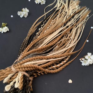 May include: A collection of braided hair extensions in shades of brown, blonde, and black. The braids are bound with a light brown string. White flowers and pearls are scattered around the braids on a dark surface.