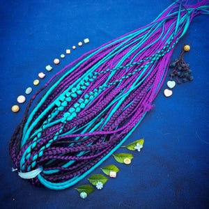 May include: A bundle of purple and teal synthetic dreadlocks, some braided, others loose, against a dark blue background. Decorative elements include small beads, leaves, and heart-shaped accents. The dreadlocks are tied together with a white band.