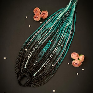 Puede incluir: Un conjunto de rastas negras, turquesas y grises, adornadas con cuentas, plumas y detalles plateados. Las rastas están dispuestas sobre una superficie negra, con rosas color melocotón y perlas dispersas.