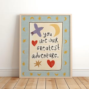 May include: A framed print featuring a light blue border with yellow stars and crescent moons. The center panel is cream-colored with the text 'you are our greatest adventure' in black, accompanied by a purple bird, two red hearts, and a yellow moon.