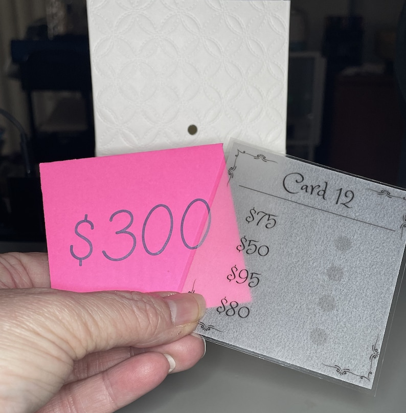 May include: A pink sticky note with the price $300 written on it, partially covering a clear plastic card with the text "Card 12" and a list of prices: $75, $50, $95, $80.