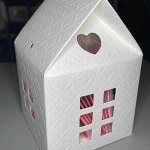 May include: White cardboard house-shaped box with a heart-shaped cutout on the roof and four rectangular cutouts on the front, revealing pink and red paper inside.