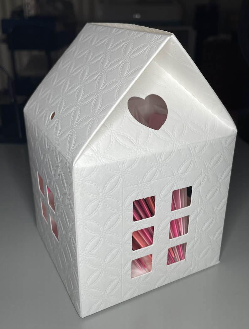 May include: White cardboard house-shaped box with a heart-shaped cutout on the roof and four rectangular cutouts on the front, revealing pink and red paper inside.