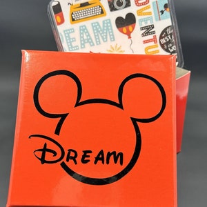 May include: Red box with a black Mickey Mouse silhouette and the word "DREAM" in a stylized font. The box is open and a small card with a colorful pattern is visible inside. The card has images of a typewriter, camera, and a Mickey Mouse balloon.