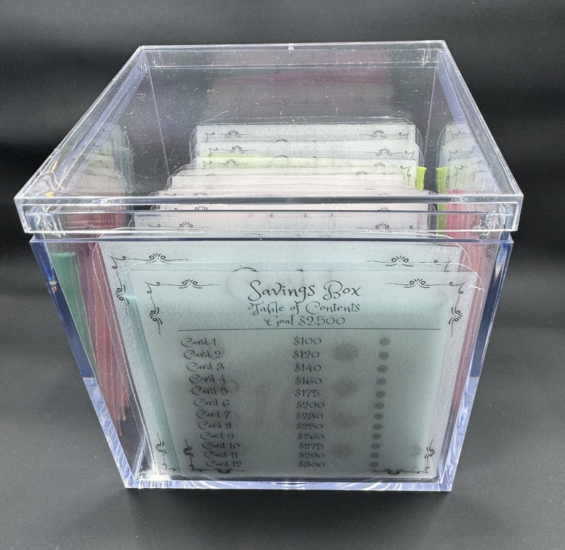 May include: A clear plastic box with a lid containing a stack of cards. The cards are labeled with numbers and dollar amounts. The text on the cards reads "Savings Box Table of Contents Goal $2,500".