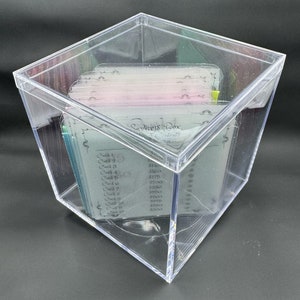 May include: A clear plastic box with a lid, containing a stack of cards with handwritten text and numbers. The text on the cards reads "Savings Box" and lists "Card 1" through "Card 12" with corresponding dollar amounts.