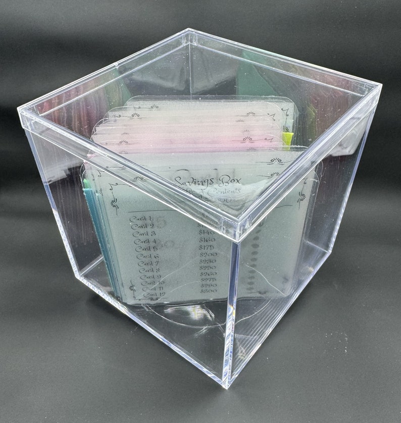 May include: A clear plastic box with a lid, containing a stack of cards with handwritten text and numbers. The text on the cards reads "Savings Box" and lists "Card 1" through "Card 12" with corresponding dollar amounts.